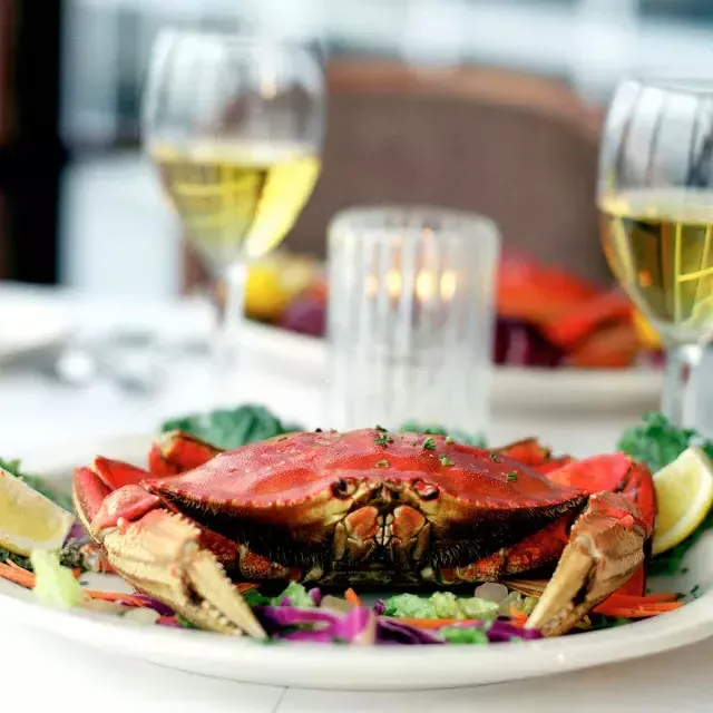 Un cangrejo Dungeness reposa en un plato en un restaurante con dos copas de vino blanco de fondo.